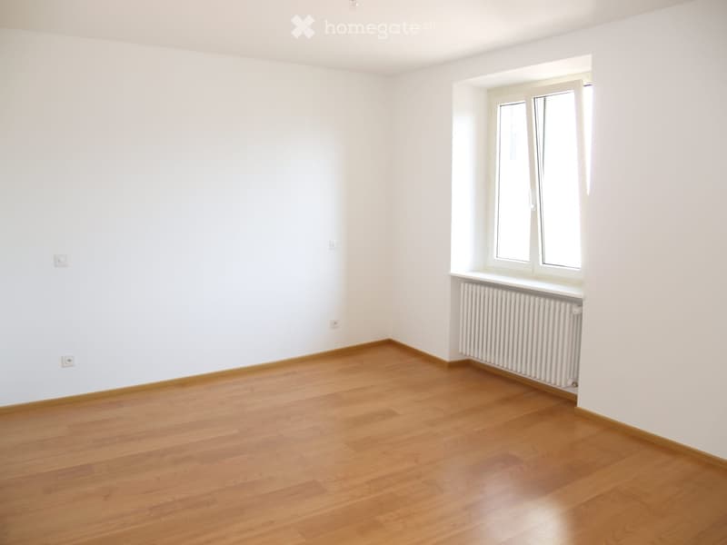Empty room with light wooden flooring, white walls, and a window with white frames and radiator below it.