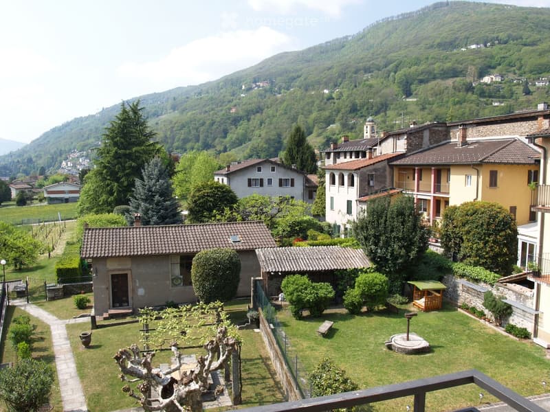 A serene garden with a well-maintained lawn, trimmed bushes, and a small wooden playhouse. The garden is bordered by a stone wall and features a small fountain and a tree pruned into an artistic shape. In the background, traditional stone houses and lush