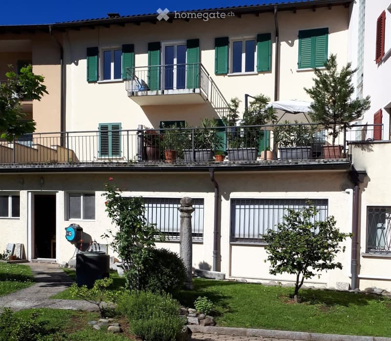 Two-story house with light yellow walls, green shutters, and multiple balconies. The ground floor has a small garden with a stone pillar, a water tank, and a hose reel. The upper floor features potted plants on the balcony and a large umbrella providing s