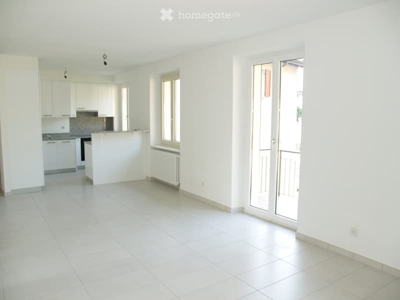 Empty room with white tiled flooring, white walls, and a sliding glass door leading to a balcony. The room is connected to a modern kitchen area with white cabinets and a countertop.