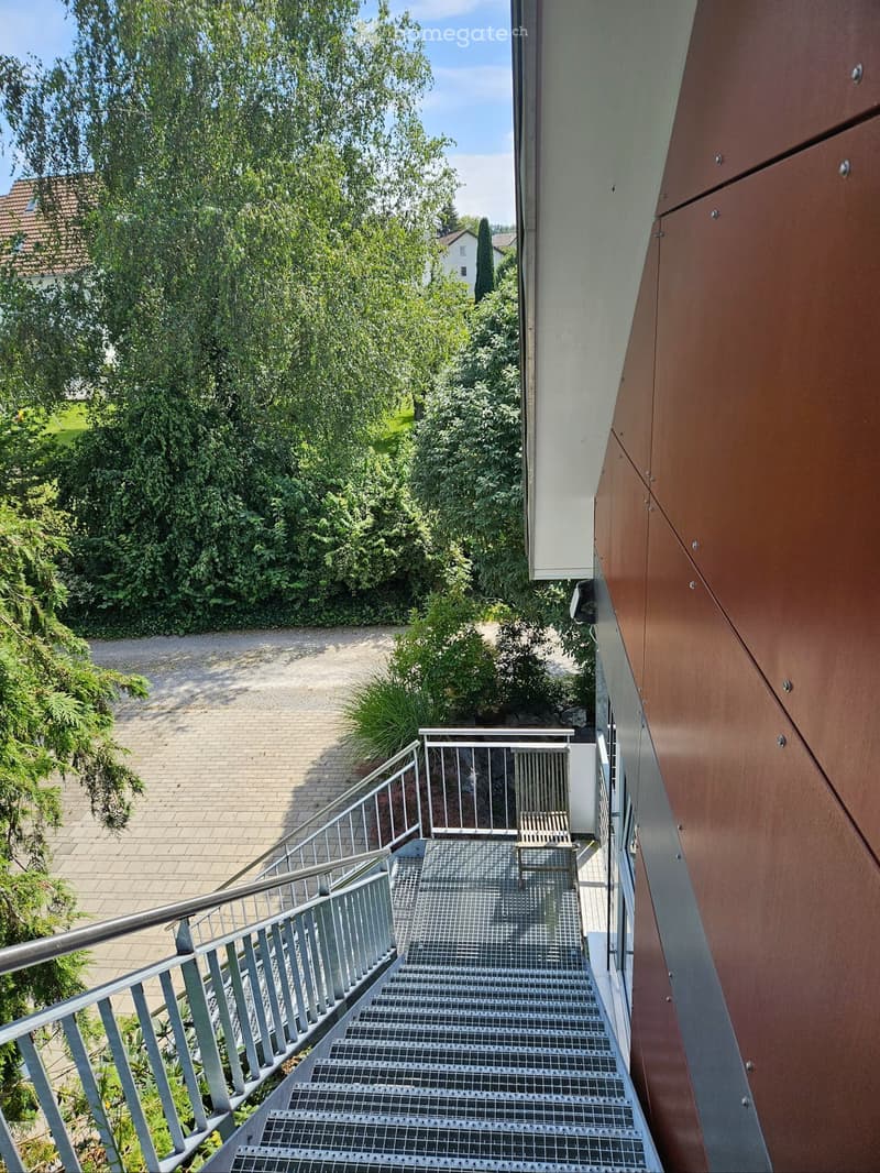 Modern staircase leading up to a balcony or terrace area with metal railings, surrounded by greenery and a paved pathway.