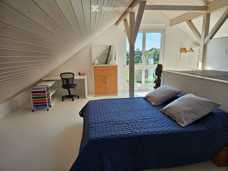 Modern attic bedroom with slanted wooden ceilings, large windows, a bed with blue bedding, a desk with a chair, a cabinet, and a small shelf with colorful storage bins.