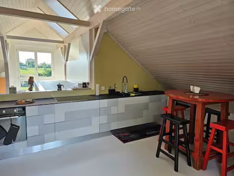Modern kitchen with grey cabinets, white countertops, and a red dining table with stools. Features a dishwasher, sink, and a view of the outdoors through large windows.