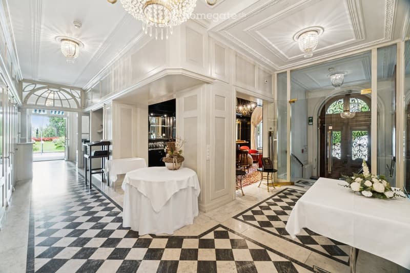 Elegant hallway with checkered black and white flooring, white walls, ornate chandeliers, and several round tables with white tablecloths, surrounded by chairs and decorative flower arrangements.