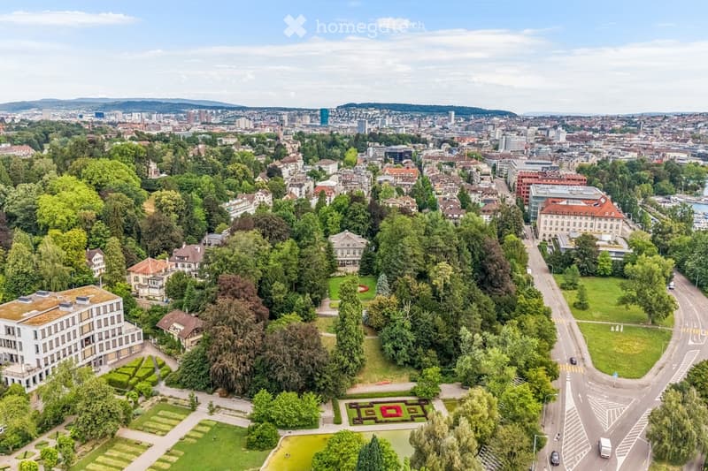 Aerial view of a cityscape with a mix of residential and commercial buildings, surrounded by green spaces and trees. The area includes a park with a pond, pathways, and a patterned garden bed.
