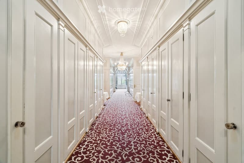 Elegant hallway with white paneled walls, ornate carpet, chandeliers, and multiple doors.