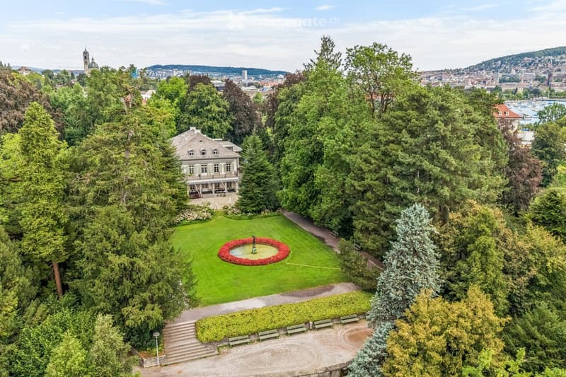 Aerial view of a lush garden area with a circular flower bed, a fountain, a house, and surrounding trees, with a cityscape in the background.