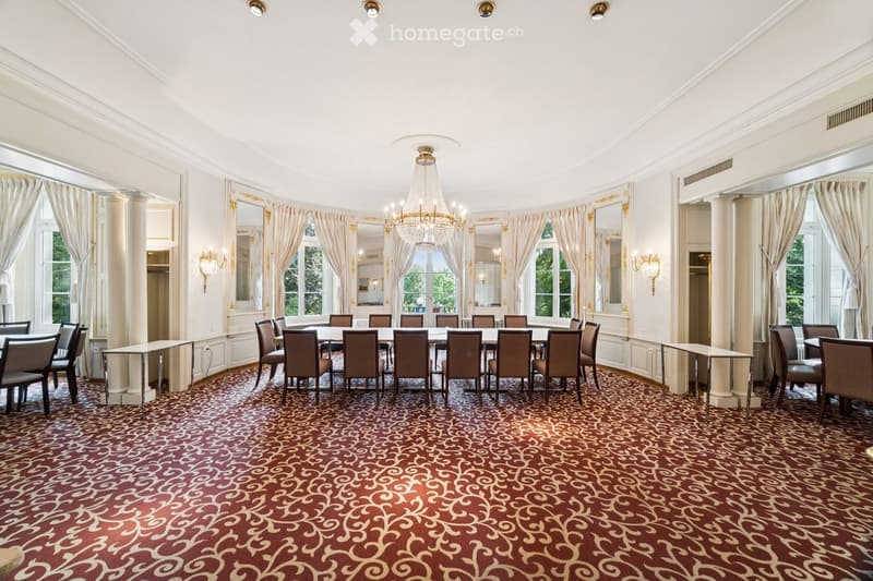 Elegant dining room with a large round table surrounded by chairs, a chandelier hanging from the ceiling, side tables, large windows with curtains, and a patterned carpet.