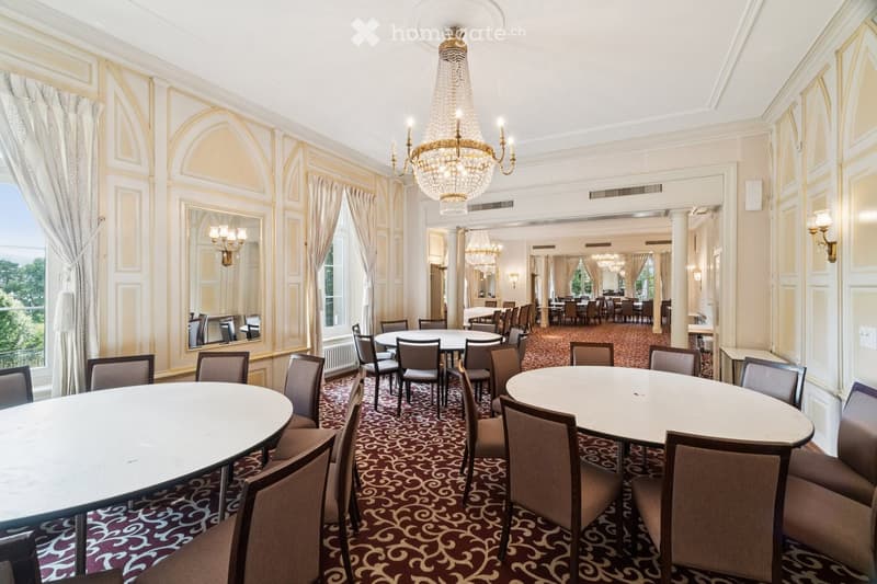 Elegant dining area with round tables, chairs, ornate chandelier, large windows with curtains, and a patterned carpet.