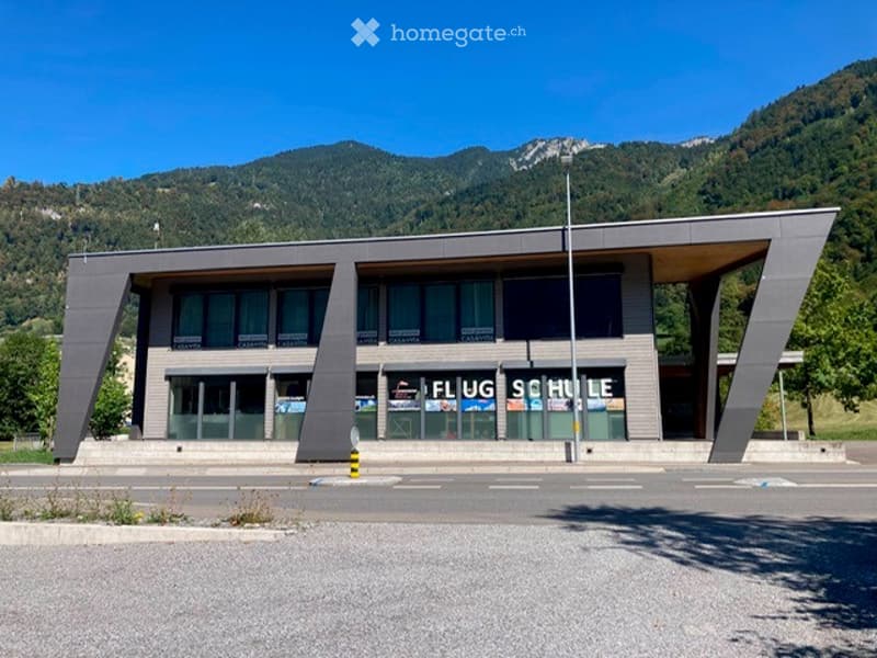 Modern building with a flat roof and large glass windows, featuring a sign that reads "FLUG SCHULE" and surrounded by a clear road and mountainous landscape.