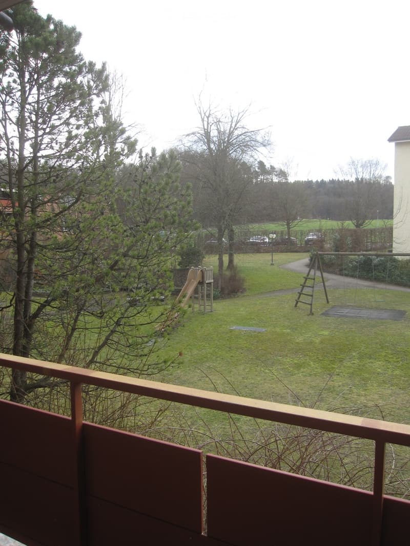 Balcony with railings, view of a grassy field with a slide and swings, trees, and a building in the distance