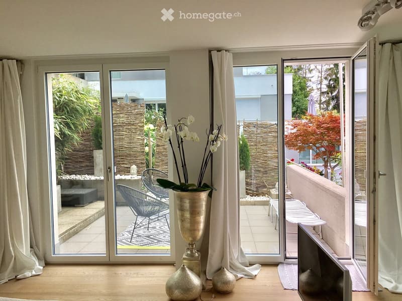 Bright room with large sliding glass doors opening to a balcony, featuring modern chairs, decorative vases, and a patterned rug.