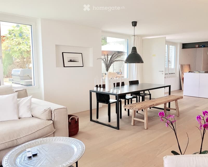 Bright, open living and dining area with light wood flooring, a beige sofa, a glass coffee table, a modern dining table with chairs and a wooden bench, and decorative vases.