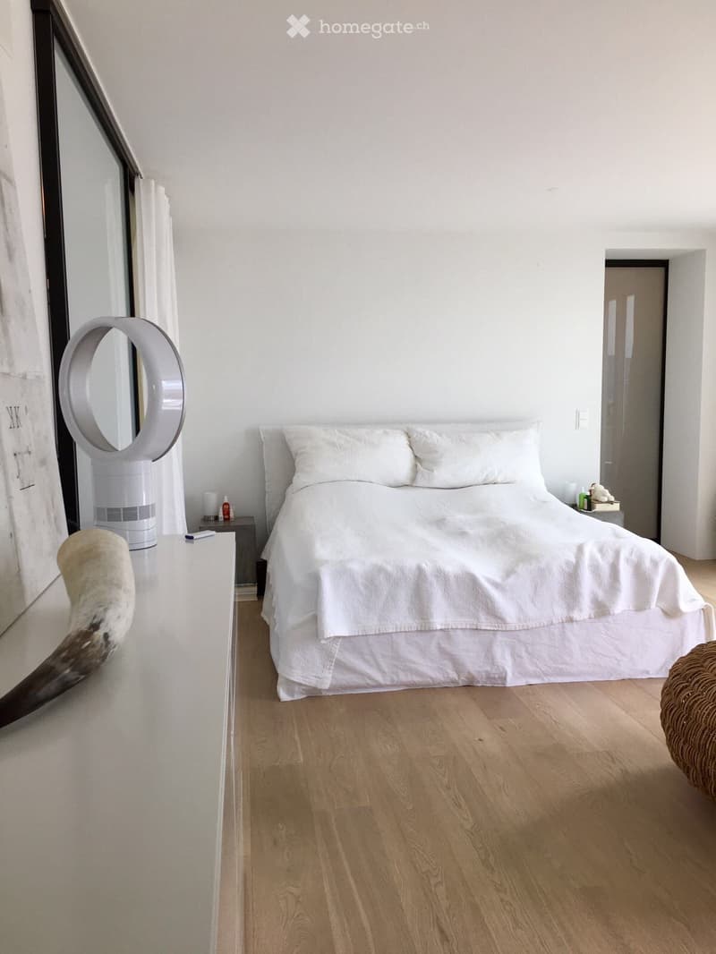 Modern bedroom with a large bed, white bedding, a nightstand, a round mirror, and a fan. The room features light wooden flooring and white walls.