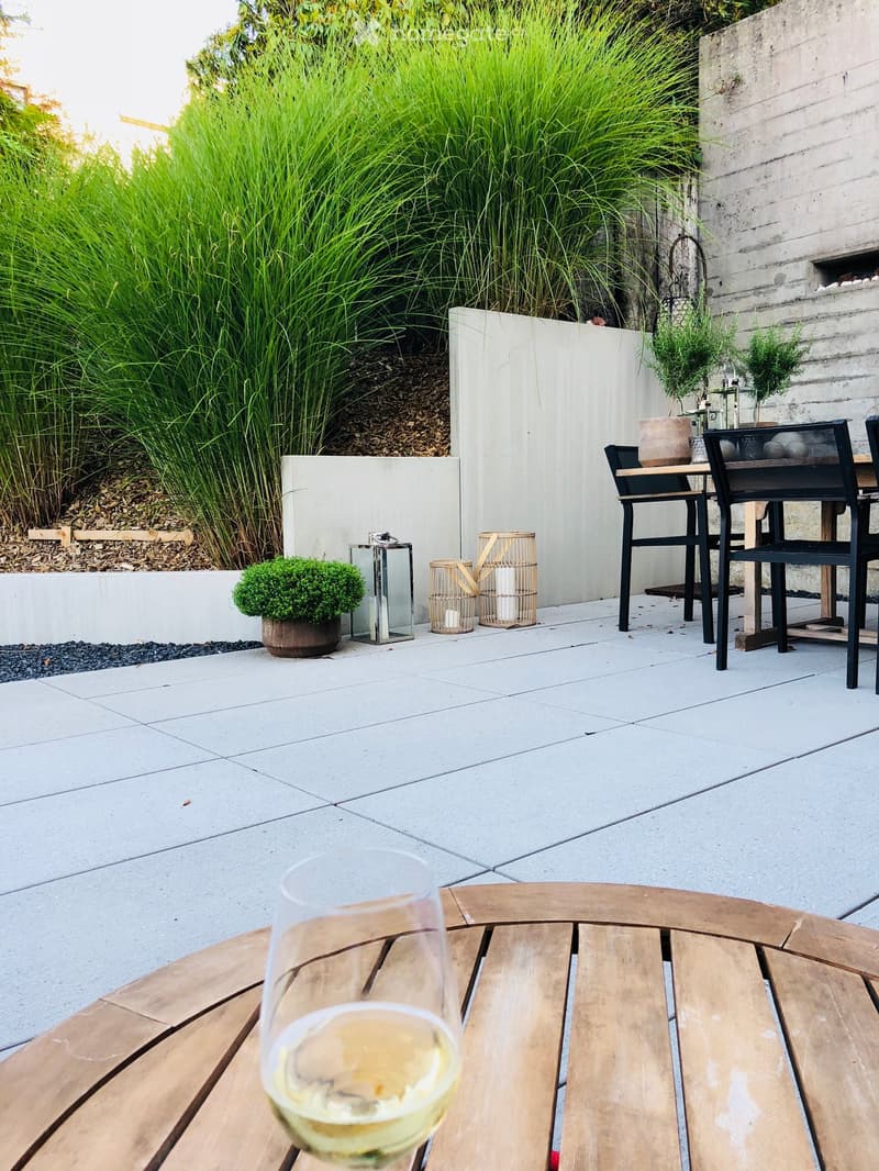 A modern outdoor patio with a wooden table, black chairs, a glass of white wine, and decorative plants including tall grasses and small shrubs.