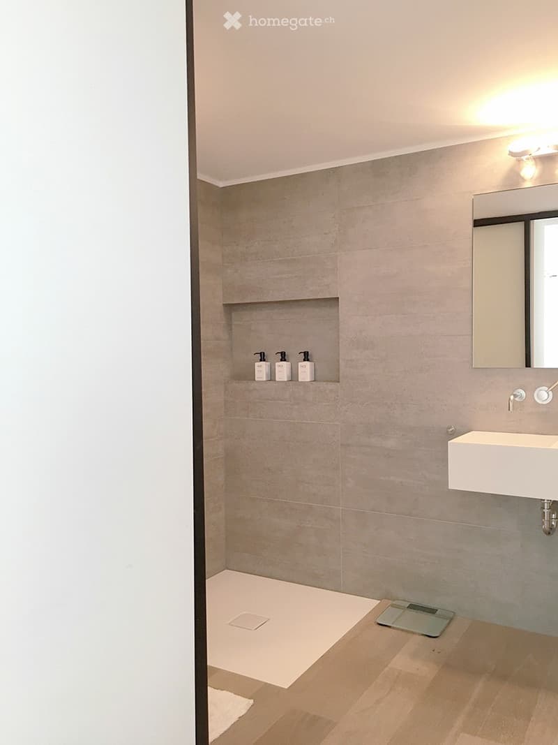 Modern bathroom with a white sink, mirror, and four bottles on a shelf. The floor is light wood, and the walls are tiled in a neutral tone.