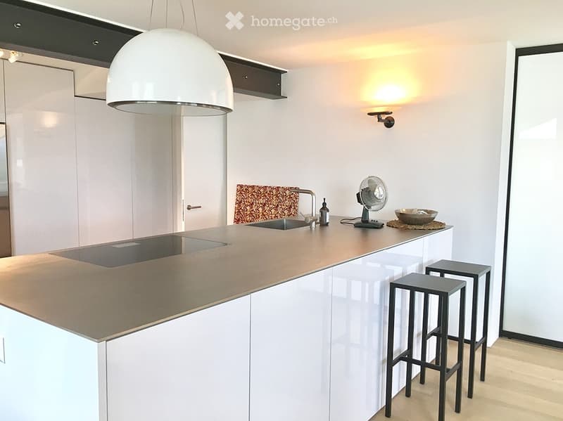 Modern kitchen with white cabinets, a central island, and a sleek countertop. Features include a hanging pendant light, two bar stools, a small fan, and a decorative bowl on the island.