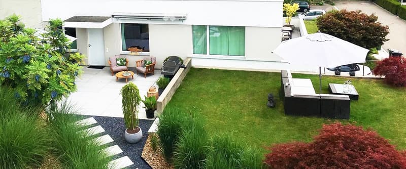 Well-maintained garden with a patio area, outdoor seating, and a small lawn. Features include a grill, potted plants, and a large umbrella for shade.