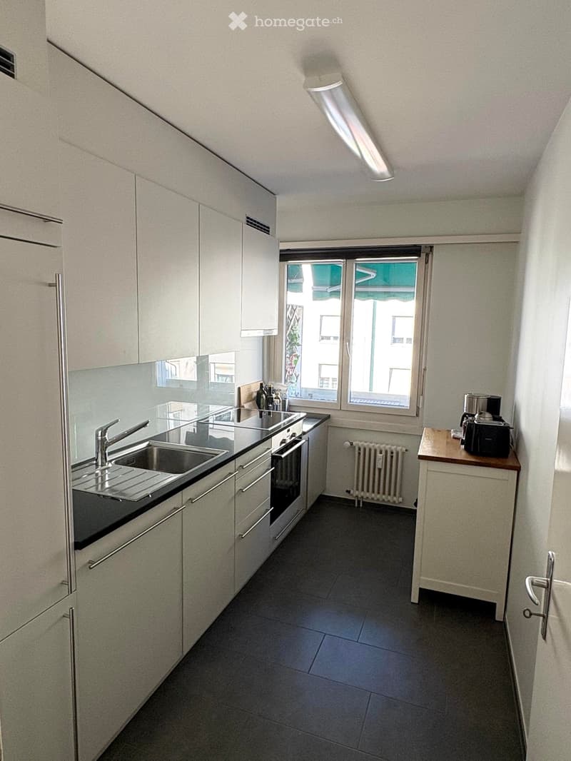 Modern kitchen with white cabinets, stainless steel sink, oven, and coffee maker on a small table. Large windows provide natural light.