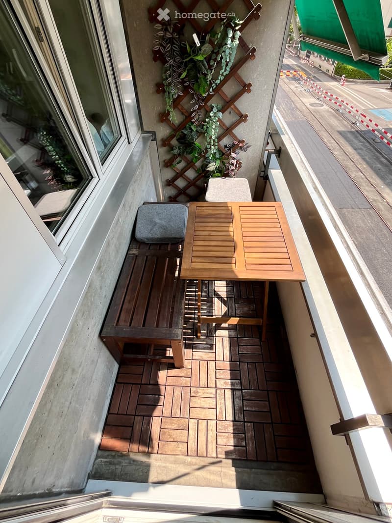 Balcony with wooden decking, a small table, two chairs, and a stool, with a decorative wooden trellis and green plants.