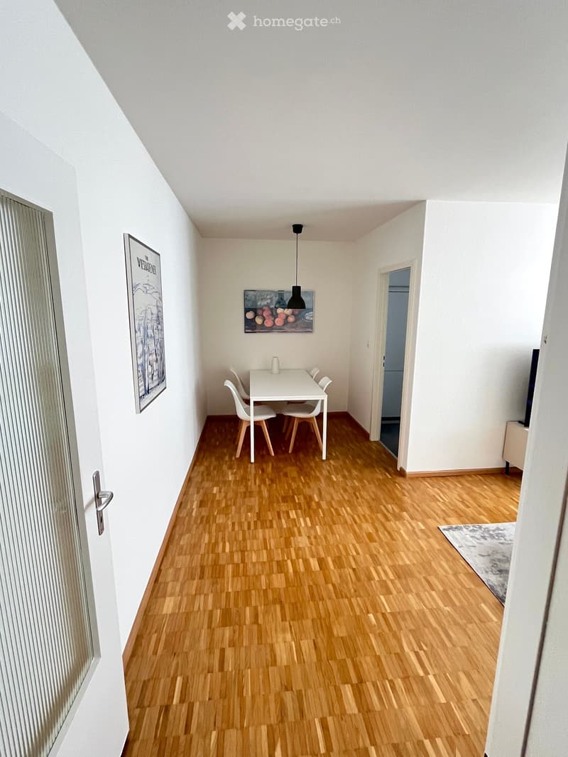 Bright, minimalist dining area with a white dining table, four white chairs, and a hanging pendant light. The room features light wooden flooring, white walls, and two picture frames on the wall.