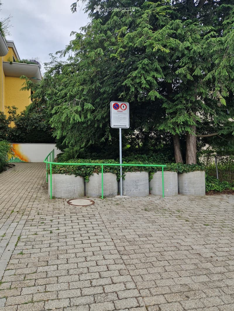 A paved outdoor area with a green metal railing, concrete planters, a signpost, and surrounding trees.