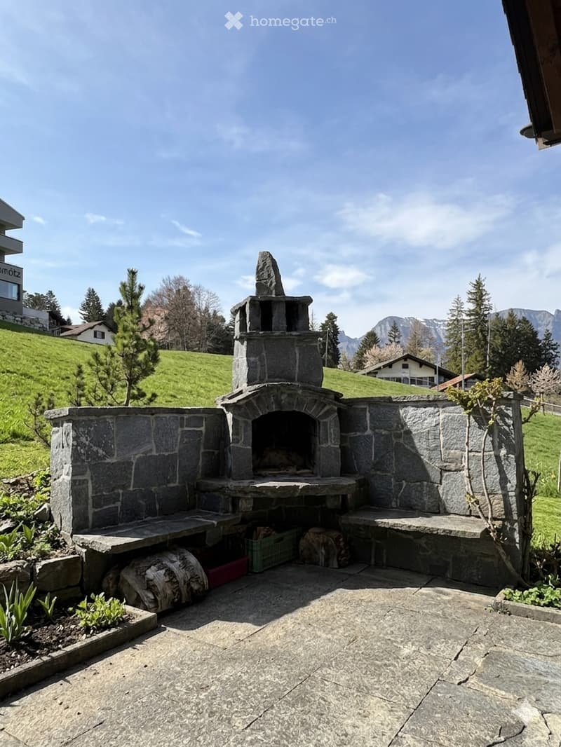 Stone fireplace with seating area, surrounded by greenery, with a view of the mountains