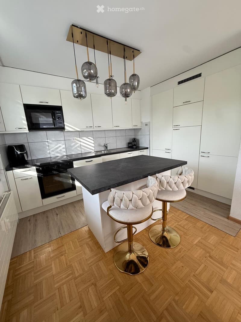 Modern kitchen with white cabinets, black countertop, and two white tufted stools with gold bases. Features include an oven, microwave, and a sink with a faucet.