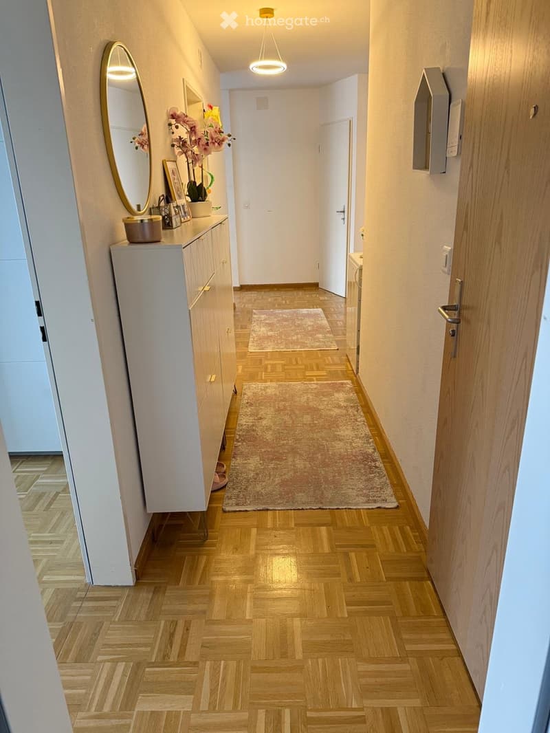 A well-lit hallway with wooden flooring, two small rugs, a small cabinet with a mirror, and decorative items on top.