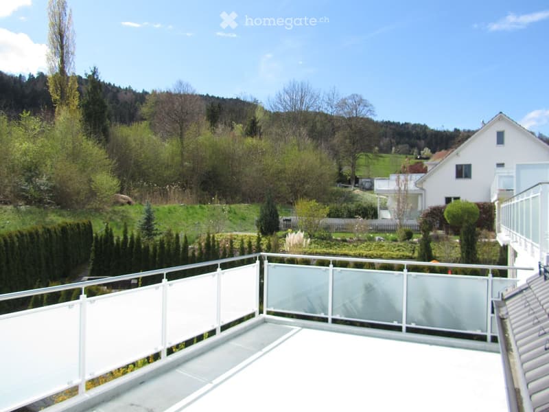 A modern balcony with a glass railing, overlooking a landscaped garden and a hilly, green landscape with trees and houses.