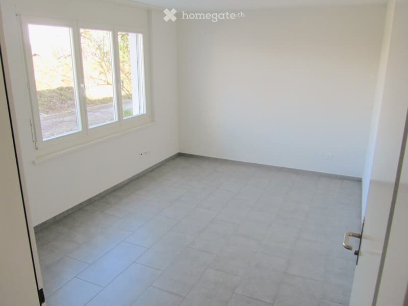 Empty room with tiled floor and white walls, featuring large windows with a view of greenery outside.
