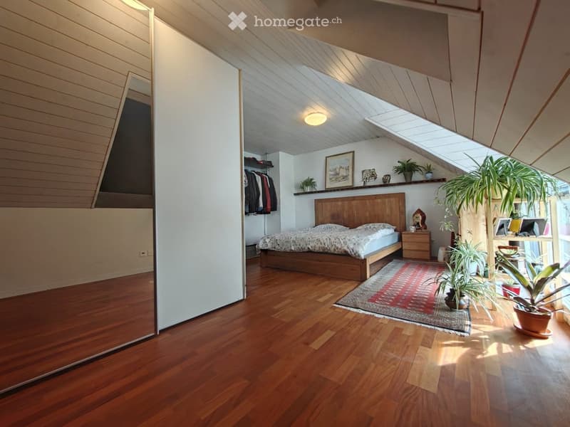 Bright, modern bedroom with wooden floors, slanted ceiling, large window, bed with wooden headboard, nightstand, potted plants, and a shelf with decorative items.