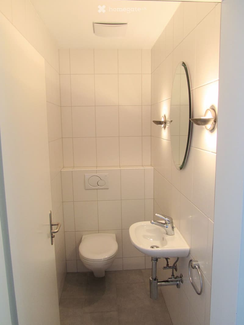 Modern bathroom with white tiled walls and floor, featuring a toilet, sink with chrome faucet, wall-mounted mirror, and built-in lighting.
