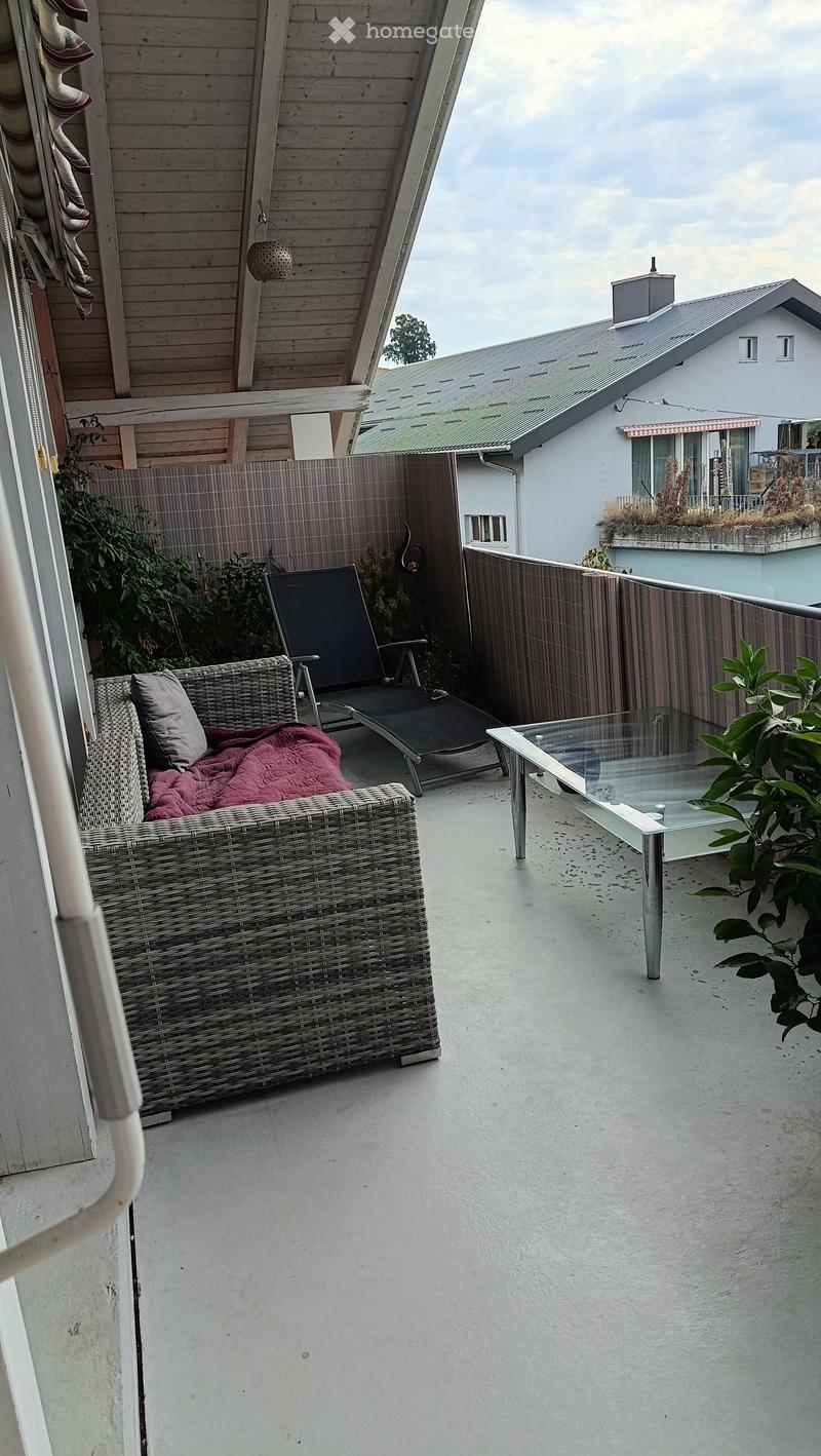 Balcony with a wicker sofa, a black lounge chair, a glass table, and a small plant. The balcony is enclosed with a wooden fence and has a view of neighboring buildings.