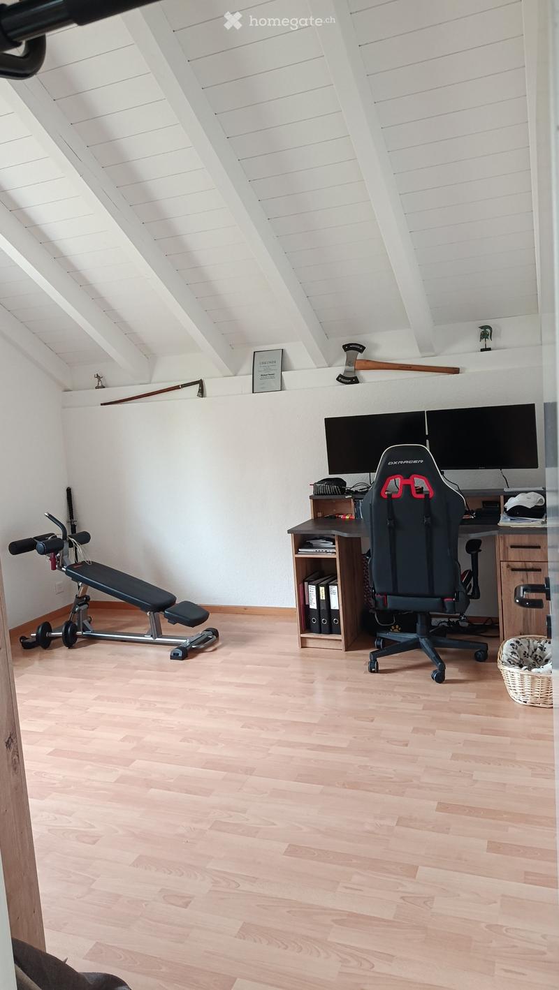 A multi-purpose room with a wooden floor, a gaming chair, desk with a screen, a small bench, and a basket. The room has a slanted white ceiling and white walls.