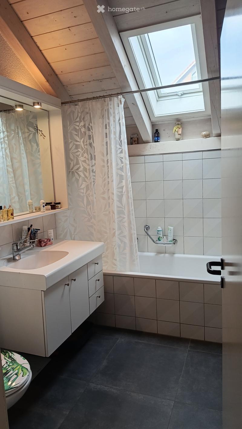 Modern bathroom with a white sink, bathtub, and mirror. Features a skylight window, dark tiled floor, and various toiletries on the sink and bathtub ledge.