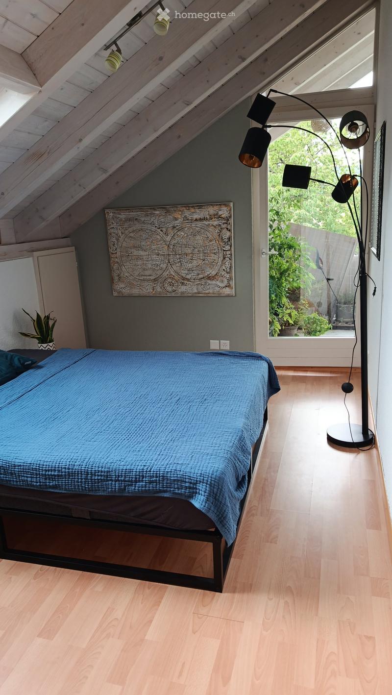 Bright attic bedroom with wooden floors, slanted ceilings, and large windows. Features a blue bedspread, a floor lamp, a potted plant, and a decorative wall art piece.