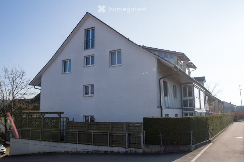 Two-story house with white exterior, multiple windows, and a balcony on the upper level. The house is surrounded by a wooden fence and a hedge, with a clear view of the street.