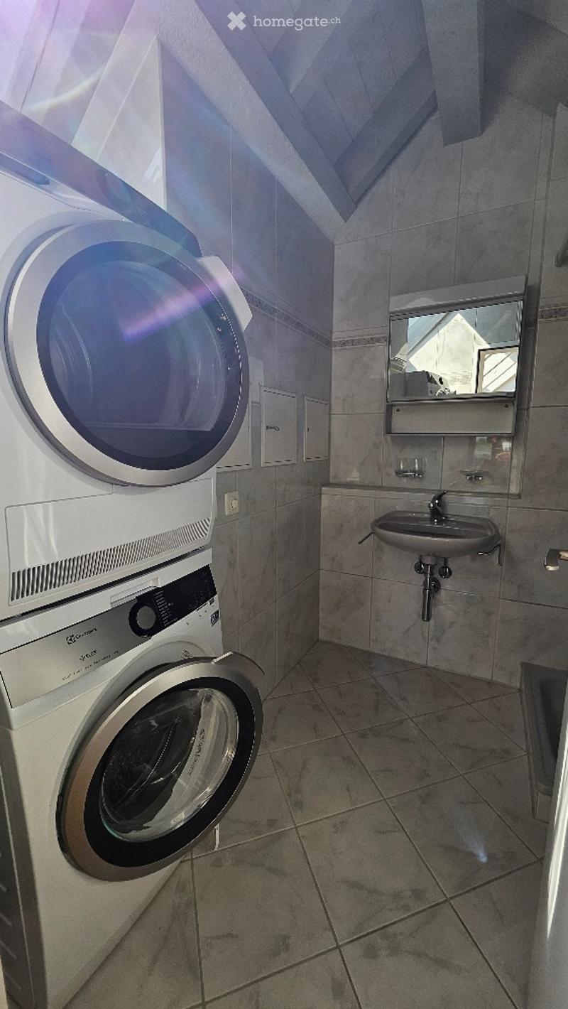 Bathroom with a washing machine, sink, and mirror. The room has tiled walls and floor, with a slanted ceiling and a small window allowing natural light.