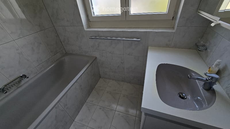 Bathroom with a bathtub, sink, and handrail. The walls and floor are tiled, and there is a window above the bathtub.