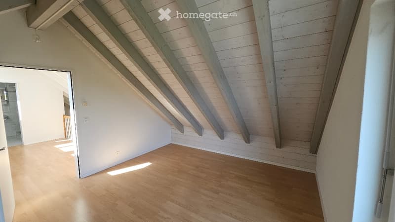 Empty room with wooden flooring and exposed wooden beams on the ceiling. Sunlight streams in from an open doorway, illuminating the space.