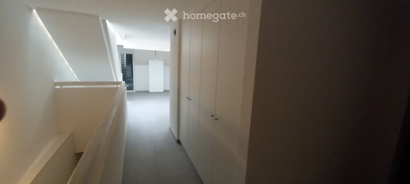 Modern, minimalist hallway with white walls, grey flooring, and a staircase leading upwards. The space is clean and uncluttered, featuring built-in storage units on one side.