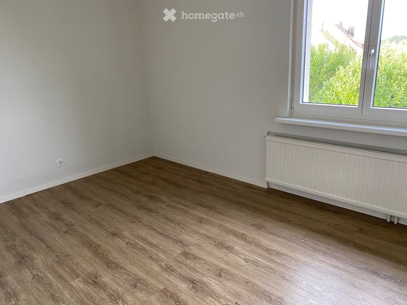 Empty room with wooden flooring, white walls, a window with white frame, and a white radiator below the window.