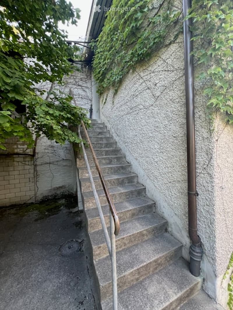 Concrete staircase with metal handrails leading up to a raised ground floor, flanked by brick and stone walls with greenery.
