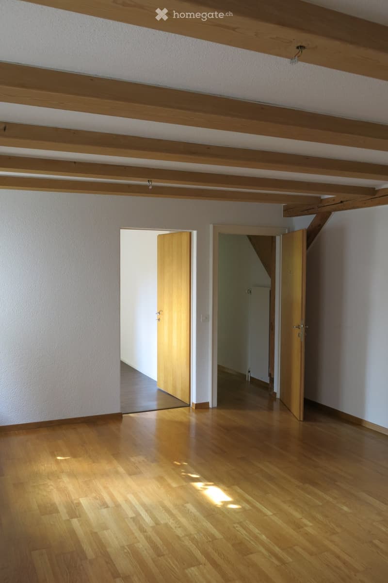 Empty room with wooden flooring and exposed wooden beams on the ceiling. Two open doors lead to adjacent spaces.