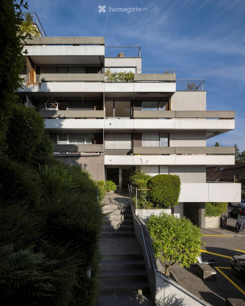 Modern three-story house with concrete balconies, large windows, and a raised ground floor accessed by stairs.