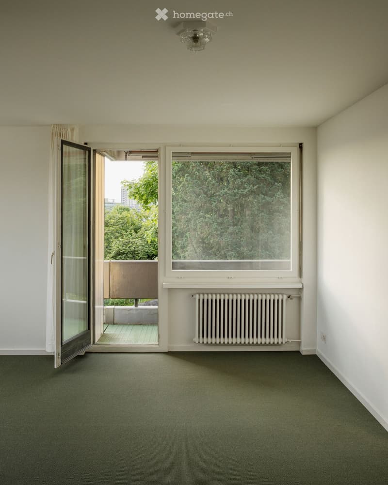 Empty room with light-colored walls, green carpet, large window with radiator below, and open sliding glass door leading to a balcony area.