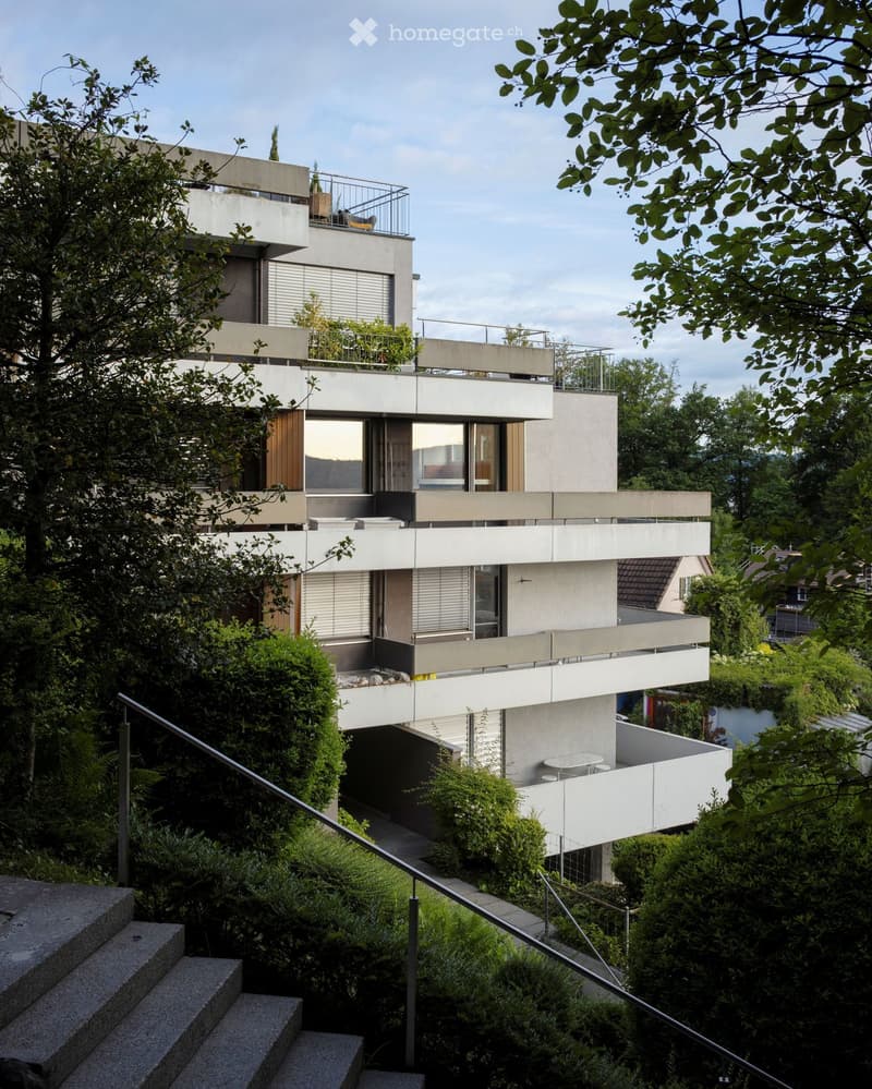 Modern multi-story house with clean lines, large windows, and balconies, surrounded by lush greenery and trees.