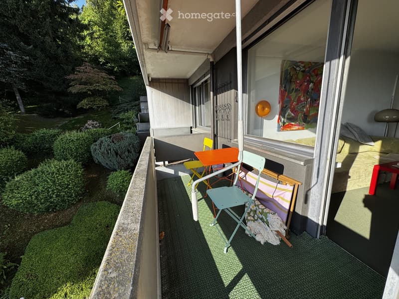 Balcony with outdoor furniture including a table, chairs, and a sofa, overlooking a green garden area.