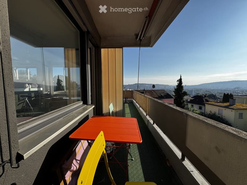 Balcony with a red table and yellow chairs, overlooking a cityscape with trees and buildings.